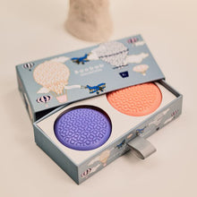 Two soaps, one purple and one orange, in a decorative box with airplane illustrations on a light background.