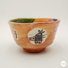 A handmade ceramic bowl with a warm orange-brown glazed exterior, a green glazed interior, and hand-painted black rabbit motifs on cream circles.