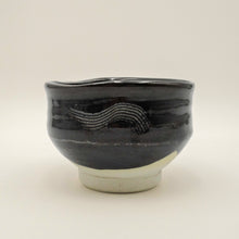 A handmade ceramic tea bowl with a glossy black glaze, featuring a textured wavy pattern on the side and a contrasting matte off-white base.