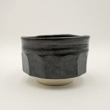 A handmade ceramic tea bowl with a dark charcoal black glaze and a speckled off-white base, photographed against a neutral light gray background.