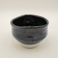 A handmade ceramic tea bowl with a glossy black glaze, featuring a textured wave pattern on the side and a contrasting white unglazed base, photographed against a neutral light gray background.