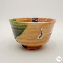 A handmade Japanese ceramic bowl with a speckled orange and yellow glaze, featuring a green glazed section on one side, a small white patch, and a dark brown center detail, shown on a neutral light gray background.