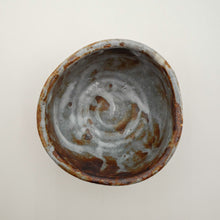 A top-down view of a handmade ceramic bowl with a rustic finish, featuring a swirling glaze pattern in gray, white, and earthy brown tones on a plain off-white background.