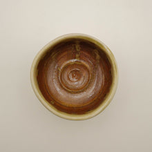 A top-down view of a handmade ceramic bowl with a glossy brown and amber glaze inside and a light beige rim, set against a plain off-white background.