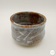 A handmade ceramic tea cup with a textured gray and brown glaze, featuring diagonal white scratch-like patterns on the front, photographed on a neutral off-white background.