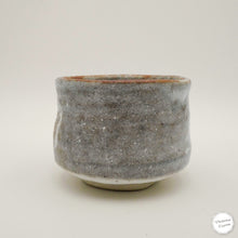 A handmade ceramic tea bowl with a textured, speckled gray and white glaze and a rustic terracotta-colored rim, photographed on a neutral light gray background.