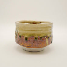 A handmade ceramic tea bowl with a glossy glaze, featuring a cream-colored top section with dripping brown and orange patterns, and a light gray base, set against a plain off-white background.