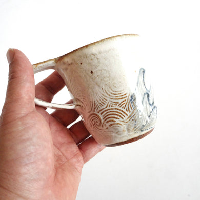 Hand holding Spako Clay mug. Blue line drawing of waves and emboss pattern, white glaze