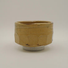 A handmade ceramic tea bowl with a textured, faceted design and a warm ochre-yellow glaze, set against a plain light gray background.
