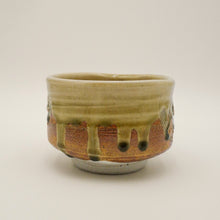 A handmade ceramic tea bowl with a textured glaze, featuring a light olive-green top section with dripping glaze over a terracotta-brown lower section, set against a plain light gray background.