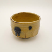 A handmade ceramic tea bowl with a textured, ribbed exterior and a glossy, warm yellow glaze, decorated with dark blue-black and brown organic drips on the side, set against a plain off-white background.
