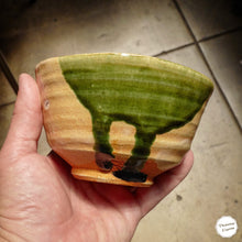 A handmade ceramic bowl with a green glaze and a ribbed clay body, held in a person's hand against a tiled floor background.