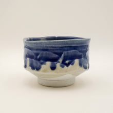 A handmade ceramic tea bowl with a blue and white glaze, featuring a textured dripping pattern on the upper half and a solid white base, photographed against a neutral off-white background.