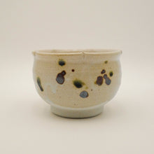 A handmade ceramic tea bowl with a speckled glaze, featuring dark brown and green spots on a light beige base, set against a plain light gray background.