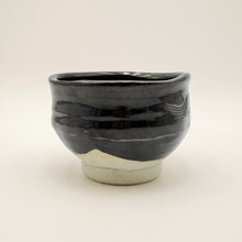 A handmade ceramic tea bowl with a glossy black glaze and a textured, unglazed light beige base, photographed against a neutral off-white background.