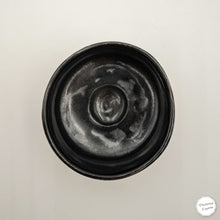 A top-down view of a round black ceramic bowl with a textured, hand-painted finish, set against a plain off-white background.