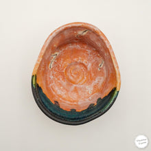 A handmade ceramic bowl with a rustic, hand-painted finish, featuring a terracotta orange interior, a dark teal-green glazed rim, and a black outer edge, shown from a top-down perspective on a light off-white background.