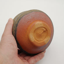 A handmade ceramic bowl with a textured glaze, held in a person's hand against a plain white background. The bowl features a warm reddish-brown center, a dark brown outer rim, and a lighter beige center.