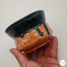 A handmade ceramic bowl with a glossy dark teal glaze dripping over a terracotta orange base, held in a person's hand against a plain light background.