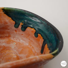 Close-up of a handmade ceramic bowl showing a glossy orange-brown interior with a dark teal and black dripping glaze along the rim, on a light gray background.
