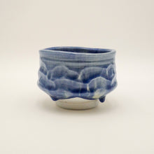 A handmade ceramic tea bowl with a textured, organic shape, featuring a glossy cobalt blue glaze and a contrasting matte off-white base, set against a plain light background.