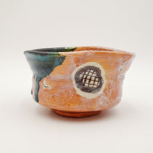 A handmade ceramic bowl with a textured terracotta orange body, featuring a dark green glazed interior, a green drip on the left side, and a circular pattern with a grid design on the front.