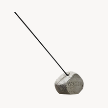 A minimalist concrete incense holder with a textured gray stone base and a single black incense stick inserted, set against a solid off-white background.