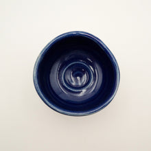 A top-down view of a small, round ceramic bowl with a glossy deep blue glaze, set against a plain off-white background.