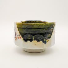 A handmade ceramic tea cup with a glossy green and black drip glaze over a beige stoneware base, photographed against a plain white background.