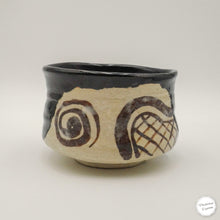 A handmade ceramic matcha bowl with a black glazed upper half and a textured beige lower half decorated with dark brown abstract swirls and a grid pattern, set against a plain light gray background.