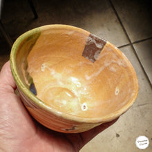 A handmade ceramic bowl with a warm orange and yellow glaze, held in a person's hand against a tiled floor background.