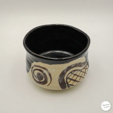 A handmade ceramic bowl with a glossy black interior and exterior glaze, decorated with a textured beige band featuring dark brown abstract swirls and a grid pattern, set against a plain light gray background.
