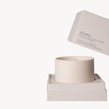A minimalist soy wax candle in a matte off-white ceramic holder, shown with its matching textured box floating above on a clean white background.