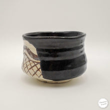A handmade ceramic tea bowl with a glossy black glaze on one half and a textured beige and brown patterned half on the other, photographed against a neutral light gray background.