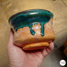 A handmade ceramic planter bowl with a glossy dark teal and orange-brown glaze, held in a person's hand against a tiled floor background.