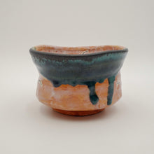 A handmade ceramic bowl with a terracotta orange base and a dark teal-blue glazed top, featuring a dripping glaze effect, set against a plain light gray background.