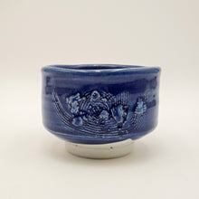 A handmade ceramic tea bowl in deep cobalt blue with a glossy glaze, featuring a raised relief wave pattern on the side and a contrasting matte off-white base.