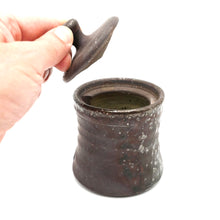 Hand holding a small brown ceramic container with a lid on a white background