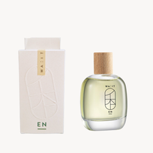 A clear glass perfume bottle with pale yellow liquid and a light wood cap stands beside a tall off-white box. The bottle and box both show the brand name WA:IT and the letters EN.
