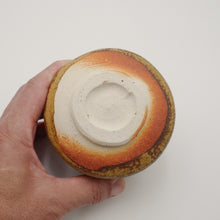 A hand holding a round ceramic bowl or vase from the bottom, showing a glazed base with concentric rings of white, orange, brown, and tan on a plain light background.