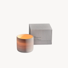 A lit cylindrical candle in a frosted ceramic jar with a warm orange glow, shown next to its minimalist light gray packaging box on a clean white background.