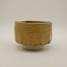 A handmade ceramic tea bowl with a textured, ribbed exterior and a scalloped pattern, featuring a warm yellow-brown glaze and a contrasting light gray base, set against a plain light gray background.