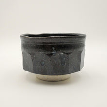 A handmade ceramic tea bowl with a dark charcoal black glaze and a small exposed beige clay foot, photographed on a neutral light gray background.