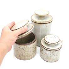 Set of three ceramic jars with speckled glaze and brown accents, one being held open.