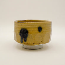 A handmade ceramic tea bowl with a textured, ribbed exterior, featuring a warm yellow-green glaze with dark brown and black ceramic drips.
