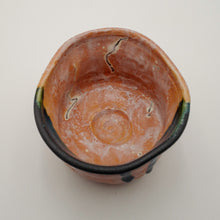 A handmade ceramic bowl with a terracotta orange interior, a black glazed rim, and subtle green and blue accents, photographed from a top-down perspective on a light off-white background.