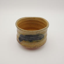 A handmade ceramic bowl with a rustic, hand-painted finish, featuring a warm tan and brown glaze with a dark charcoal/black band around the middle, set against a plain light gray background.