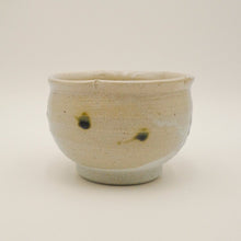 A handmade Japanese-style ceramic tea bowl with a speckled beige and off-white glaze, featuring two small dark green spots, set against a plain light background.