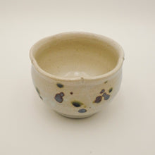 A handmade ceramic tea bowl with a speckled beige glaze and dark brown and green spots, photographed on a clean white background.