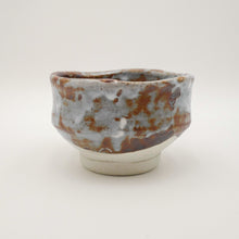 A handmade ceramic tea bowl with a rustic glaze, featuring a mottled pattern of brown, gray, and white on a light beige base, set against a plain light gray background.